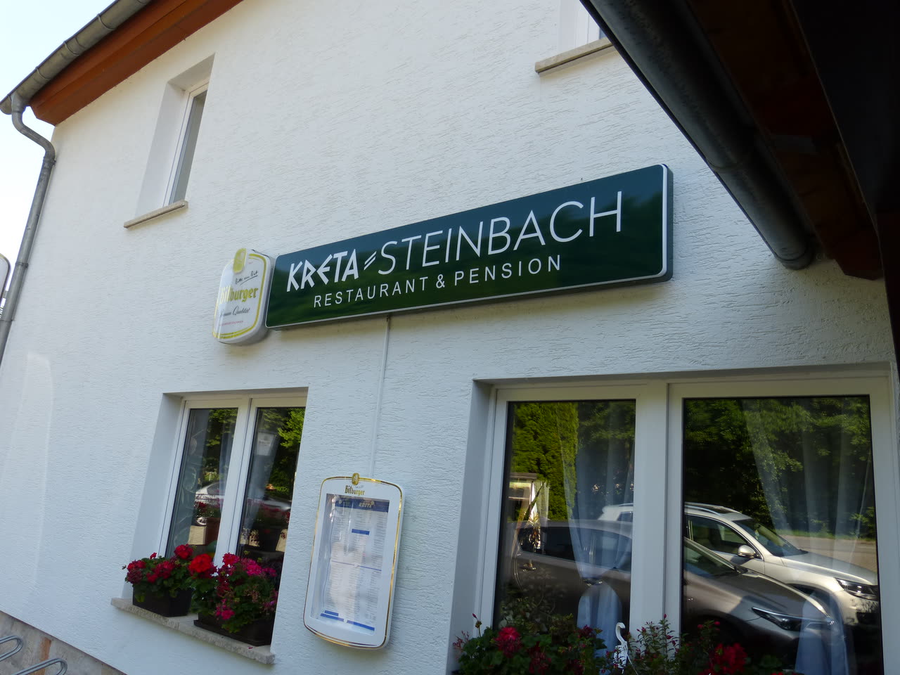 Das dunkelgrüne Firmenschild mit weißer Aufschrift Kreta Steinbach Restaurant und Pension an der weiß verputzten Hauswand