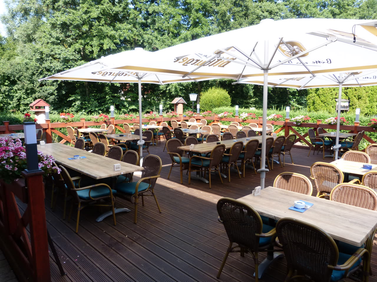 Die Sonnenterrasse mit Holzdeck, Korbstühlen unter weißen Sonnenschirmen und blühenden Geranien am Geländer